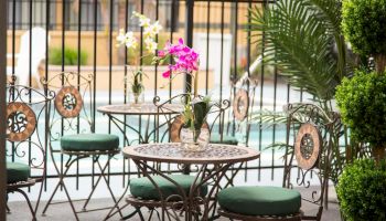 An outdoor patio area with metal chairs and tables, each adorned with green cushions and a vase of flowers, surrounded by greenery and a pool in the background.