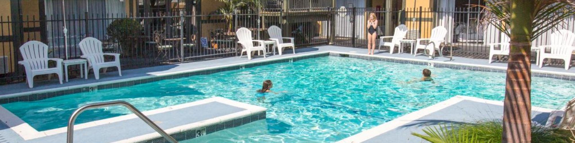 The image depicts an outdoor pool at a motel or hotel, with several people swimming and lounging, and some chairs and umbrellas surrounding the pool area.