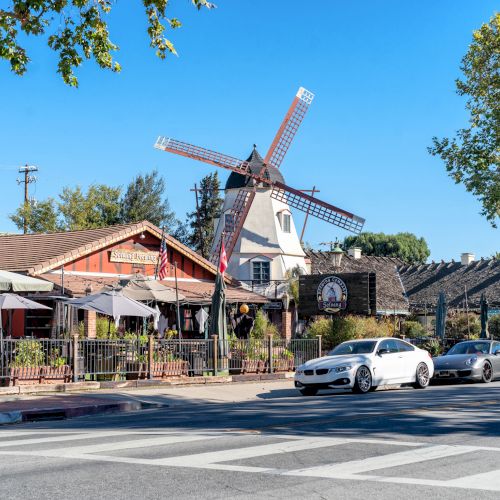 The image shows a sunny street scene with a windmill, restaurants, outdoor seating, and cars parked alongside the road.