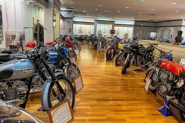 A museum room filled with a variety of vintage motorcycles displayed on a polished wooden floor with informational plaques.