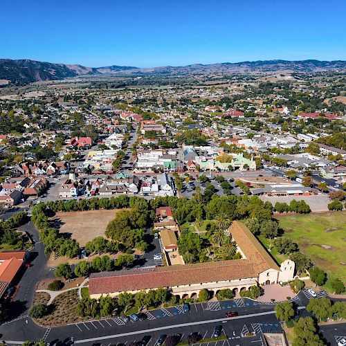 This is an aerial view of a town with buildings, streets, and open green spaces, surrounded by hills and mountains in the background.