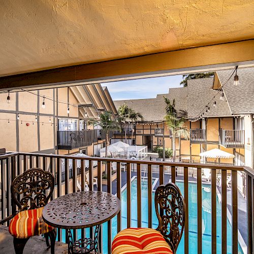 A balcony with a round table and chairs overlooks a pool area surrounded by buildings with a Tudor-style architecture.