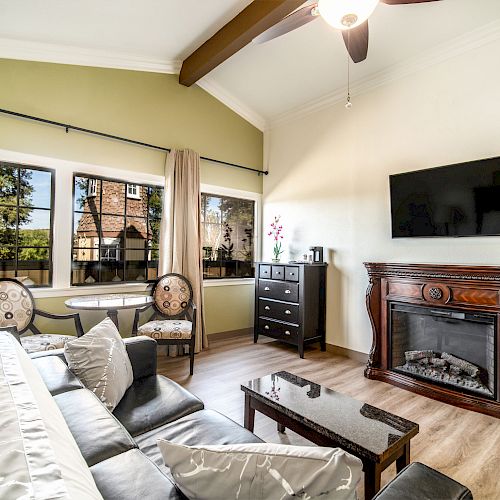 A cozy living room with a black sofa, a wall-mounted TV, a wooden fireplace, a dresser, a small table with two chairs, and large windows.