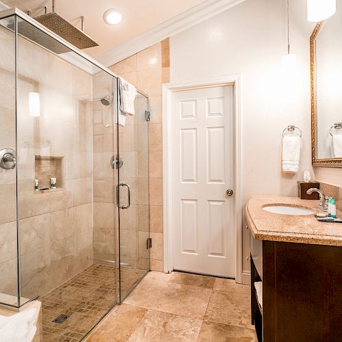 A modern bathroom with a glass-enclosed shower, a vanity with a sink, a mirror, and towel racks. The space is well-lit and tiled.