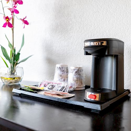 A coffee maker sits on a counter beside a vase with pink flowers. Nearby are coffee sachets, creamers, and disposable cups.