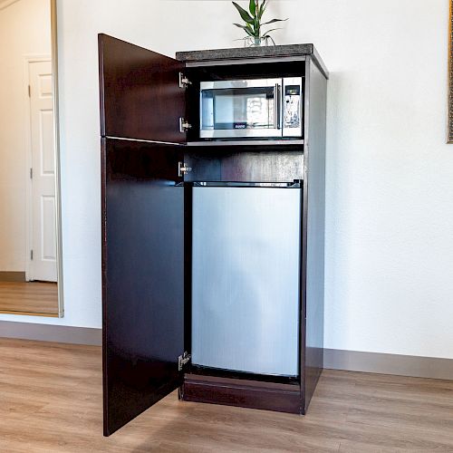 A small standing cabinet holds a microwave and a mini fridge with its doors open, located in a room with wooden floor and minimal decor.