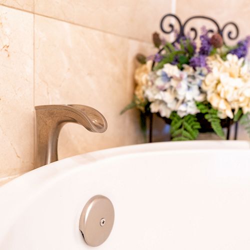 A close-up of a bathtub faucet with a floral arrangement in the background, placed against a tiled wall.