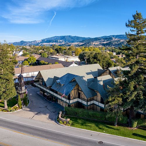 A Tudor-style building surrounded by trees, set against a backdrop of mountains and a clear blue sky, is seen from an aerial perspective.