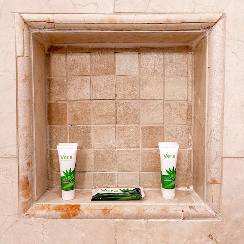 The image shows a shower niche with tiled walls, containing two tubes labeled 'Vera' and a matching sachet, all with green leaf designs, neatly displayed.