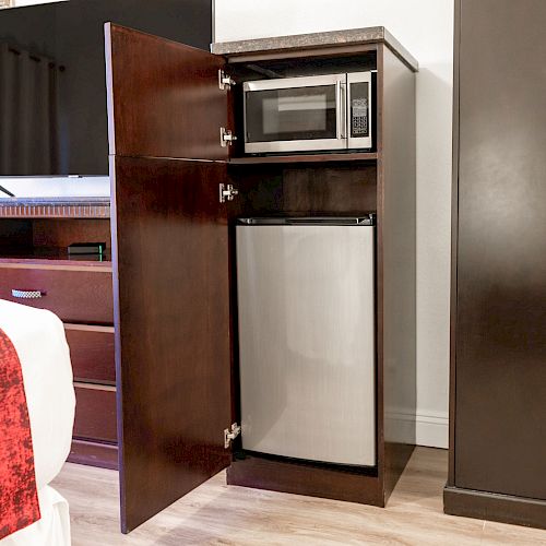 A small refrigerator and microwave are inside an open wooden cabinet in a room with a TV, dresser, and bed with a red blanket.