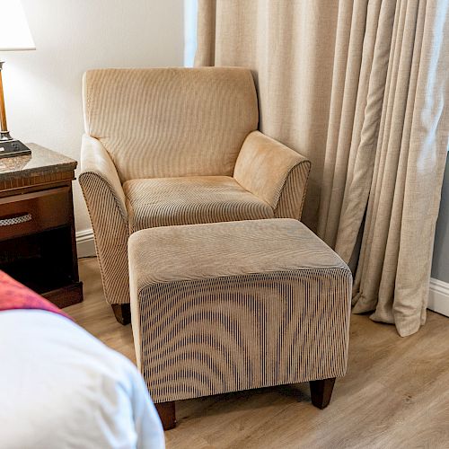 A cozy armchair with an ottoman next to a bedside table and lamp, with curtains in the background. The floor is wooden.