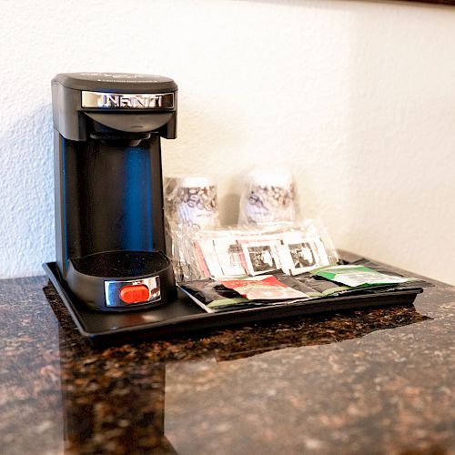 The image shows a coffee maker on a countertop, accompanied by cups and assorted coffee and tea packets arranged on a tray.