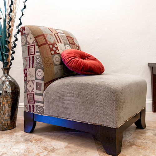 A modern living room corner with a stylish chair, red pillow, potted plant on a table, and a decorative vase with tall branches next to it.