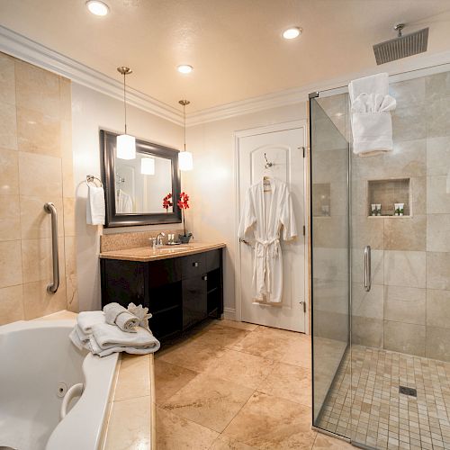 A modern bathroom with a glass shower, bathtub, vanity with sink, towels, and bathrobes hanging by the door, featuring neutral tones.