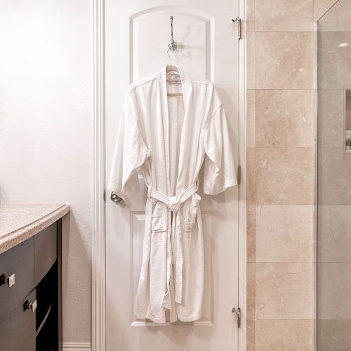 A bathroom with a countertop and sink, a red flower, a white robe hanging on a door, and a glass-enclosed shower with toiletries.