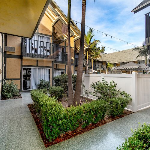 This image shows an outdoor courtyard with neatly trimmed shrubs, palm trees, and white fences, surrounded by balconies and buildings.