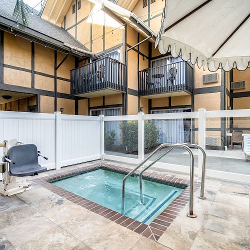 The image shows a small outdoor hot tub with a white fence, a chair lift, an umbrella, and a building with balconies in the background.