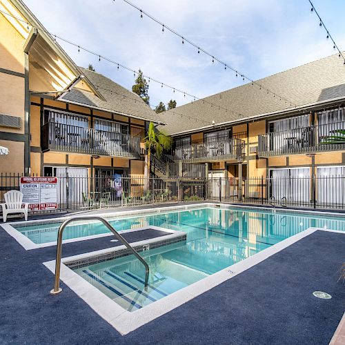 An outdoor swimming pool is surrounded by a multi-story building with balconies, chairs, and plants under string lights in the courtyard.