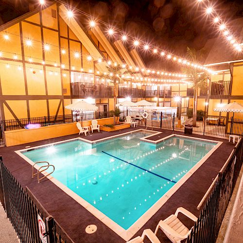 The image shows a lit-up outdoor swimming pool surrounded by a building with a yellow façade and string lights above, creating a cozy nighttime ambiance.