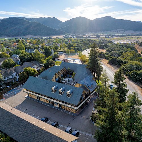 Aerial view of a suburban area with a large building in the foreground, surrounded by trees and roads, with mountains in the background.