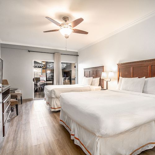 The image shows a hotel room with two large beds, a ceiling fan, a desk, and a dresser. The room has a wooden floor and modern decor.