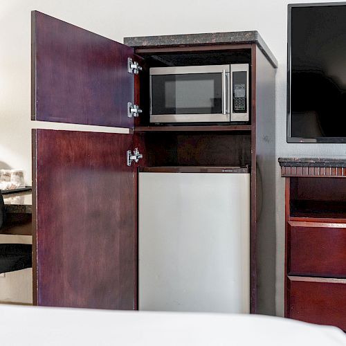 The image shows a hotel room setup with a microwave, mini fridge, TV, desk, chair, and a lamp on the left side.