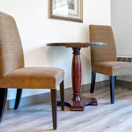 The image shows two brown upholstered chairs next to a small round table with a wooden base, in a room with wooden floors and an air conditioner.