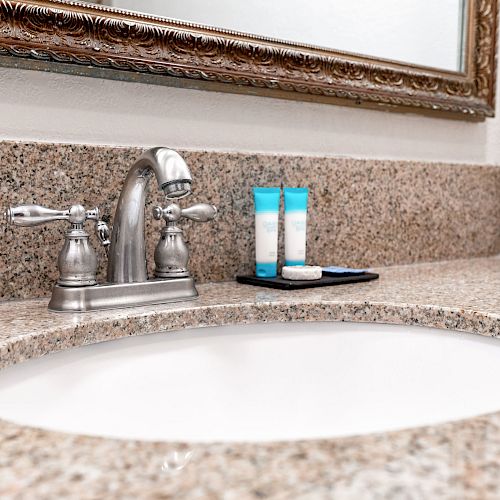 A bathroom countertop with a sink, a chrome faucet, a mirror, and toiletries including two bottles and a soap dish sitting on the right side.