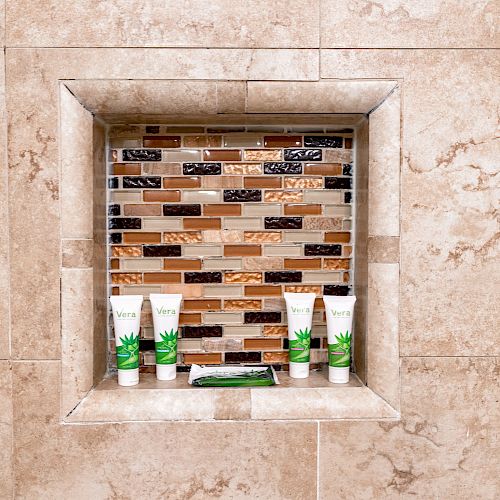 A tiled shower niche holds five bottles of toiletries against a multicolored tile background, set within beige stone tiles.