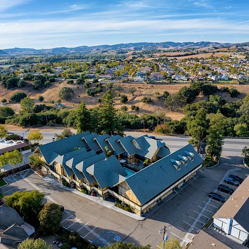 Aerial view of a building with a distinctive green roof surrounded by parking spaces, situated in a hilly suburban area with houses and greenery.