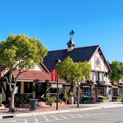 A quaint, picturesque street featuring charming buildings with pitched roofs, trees, and a clear blue sky in the background, creating a serene atmosphere.