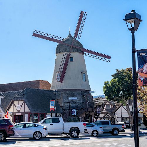 A charming street with a large windmill surrounded by European-style buildings, parked cars, and a streetlight with a banner featuring a musician.