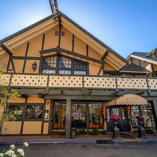 A yellow and black half-timbered building with a gable roof and balcony, featuring an awning-covered entrance and decorative potted plants.