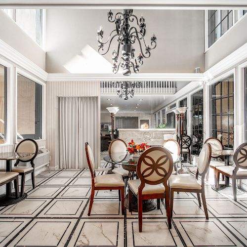 A stylish, well-lit dining area with a chandelier, an elegant table and eight chairs, surrounded by large windows and a white, patterned floor.