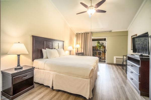 A well-lit hotel room with a king-size bed, bedside tables with lamps, a ceiling fan, a TV, and a dresser, leading to a balcony.