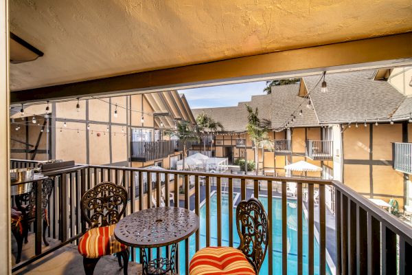 A cozy balcony with ornate table and chairs overlooks a swimming pool in a courtyard of a charming, rustic-style building.