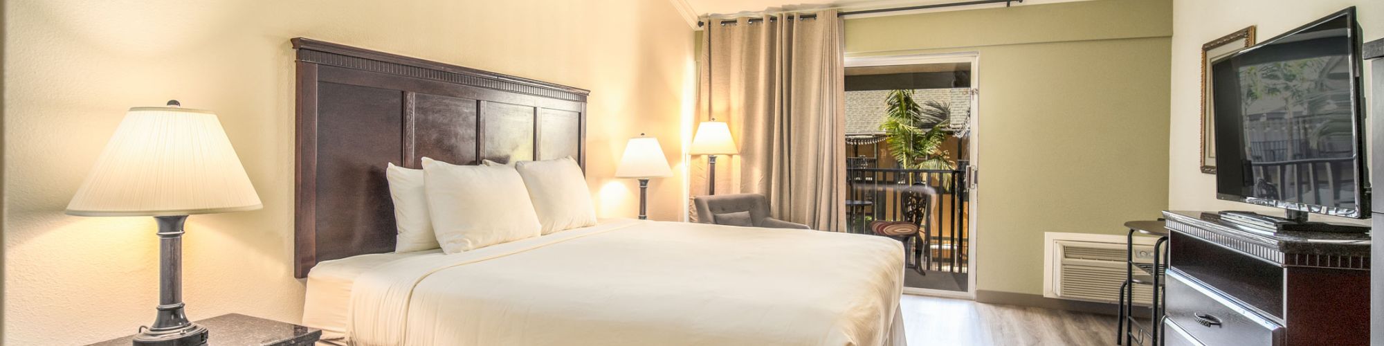 A neatly arranged hotel room with a king-sized bed, bedside tables, lamps, a ceiling fan, a dresser with a TV, and a balcony area in the background.