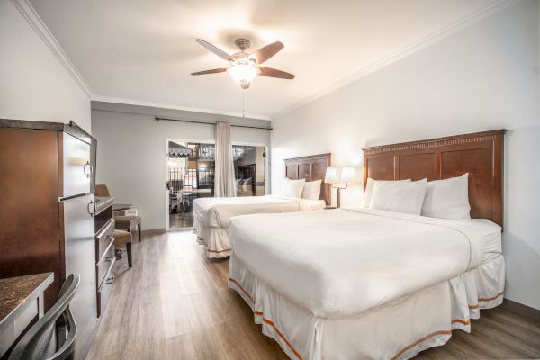 A hotel room with two queen beds, a ceiling fan, desk, chairs, and a dresser. The room is well-lit with modern decor and wooden flooring.