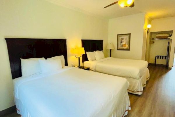 The image shows a hotel room with two neatly made double beds, a ceiling fan, a bedside lamp, and a framed picture on the wall.