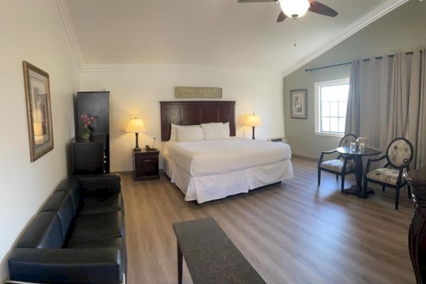 A spacious hotel room with a large bed, sofa, table, chairs, nightstands, lamps, artwork, a ceiling fan, and a window with curtains ending the sentence.