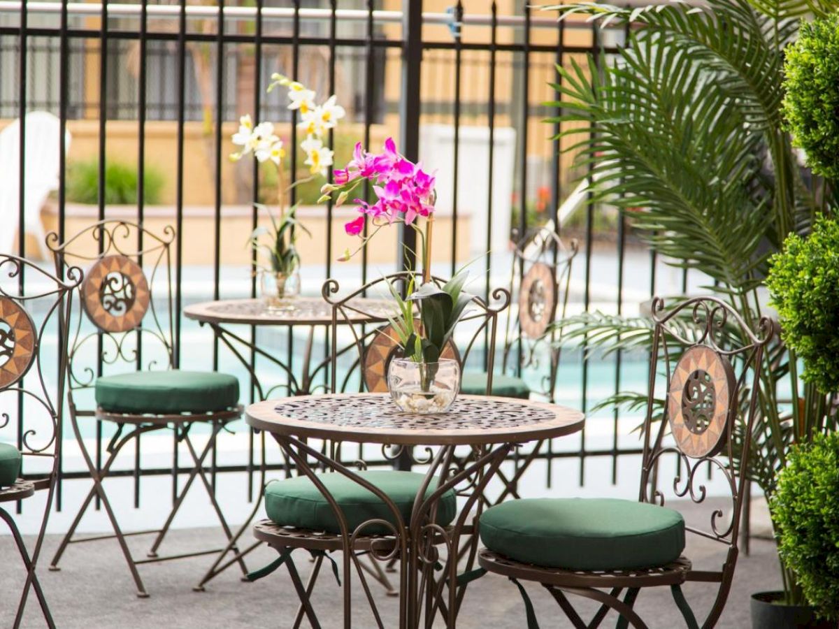 The image shows an outdoor seating area with ornate metal chairs and tables. Each table has a vase with flowers, and there's greenery around.