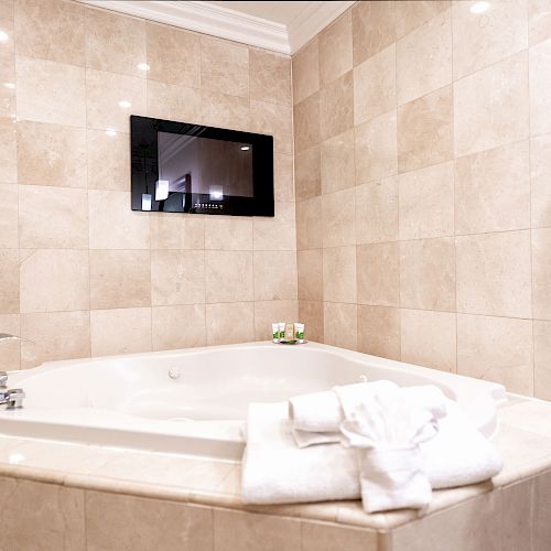 A corner bathtub with white tiles, a folded towel, and a TV screen mounted on the wall in a bathroom. The tap is on the left side.