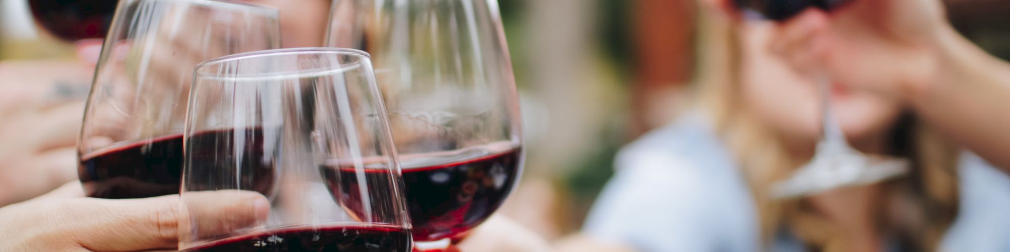 A group of people clinking glasses of red wine together in a celebratory toast, with blurred background of an outdoor setting.