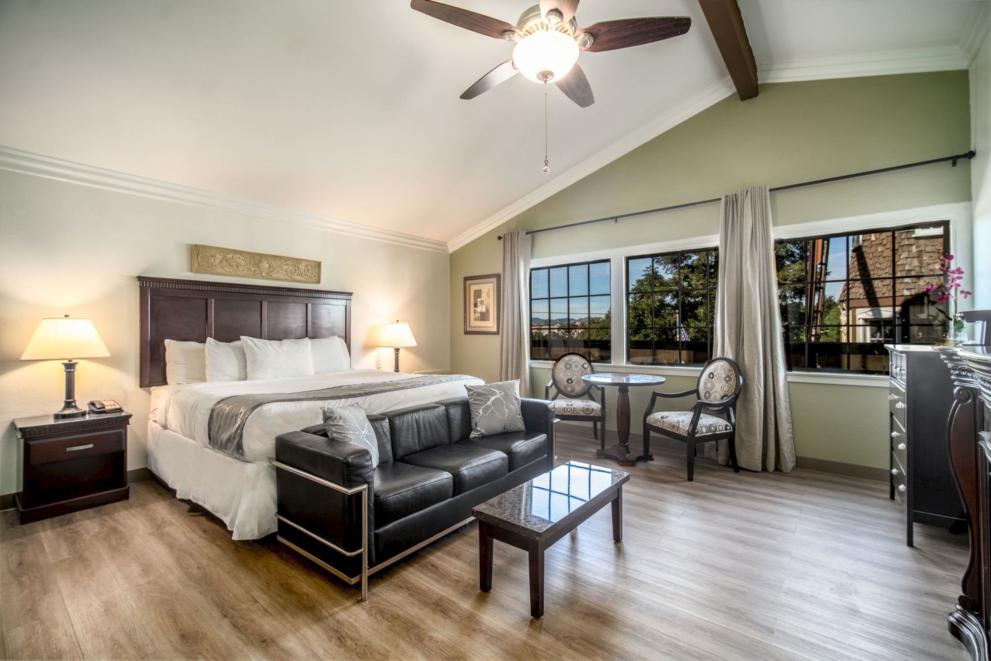 A spacious, well-lit bedroom features a king-size bed, sofa, coffee table, small dining set, and large windows with natural light streaming in.